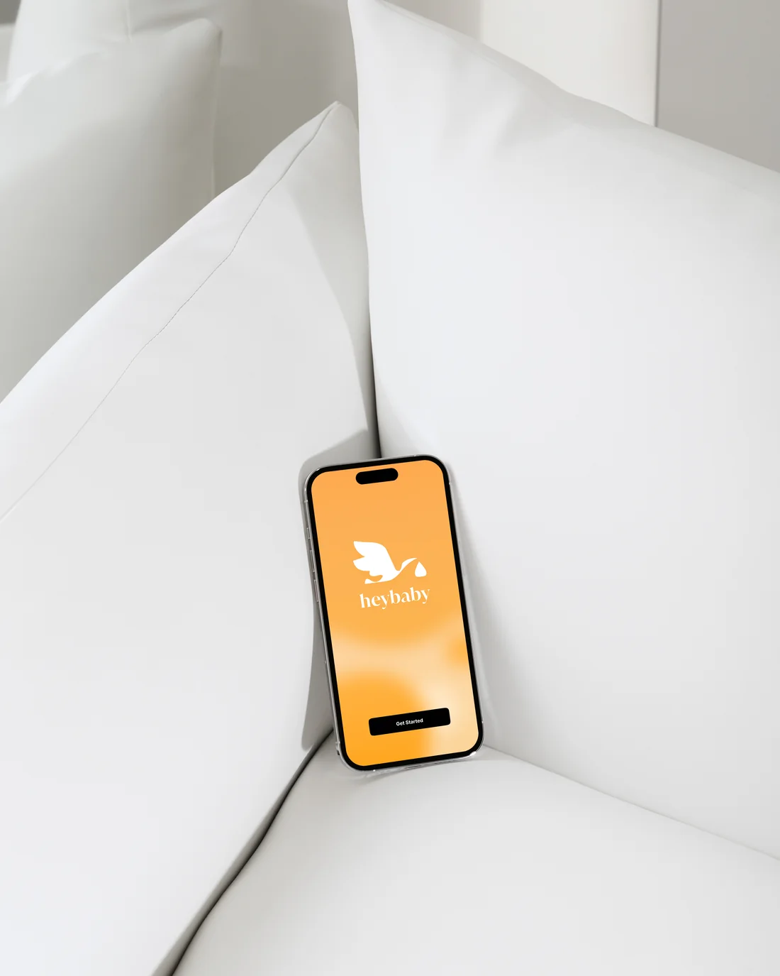 A phone resting on a white couch showing the orange HeyBaby welcome screen