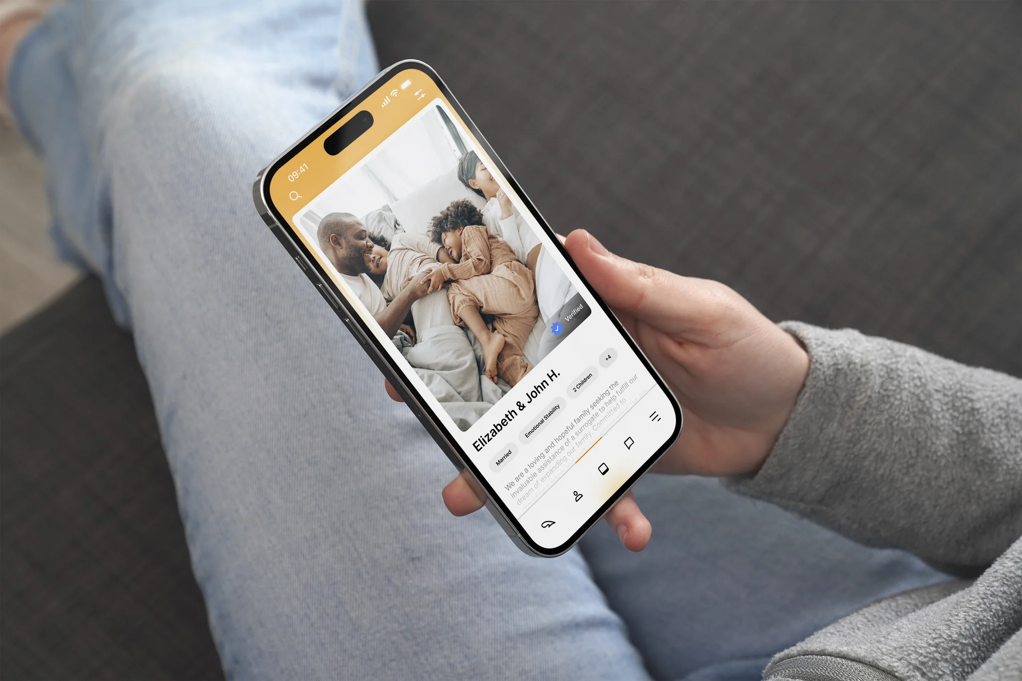 A person on a couch holding a phone showing a HeyBaby family profile